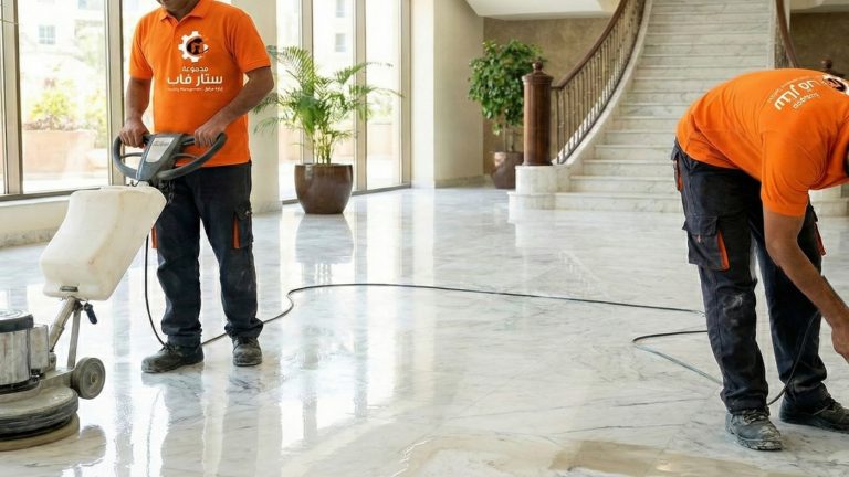 عاملان من شركة ستار فاب يرتديان زياً برتقالياً وشعار الشركة، يستخدمان ماكينة جلي وتلميع كبيرة لتنظيف أرضية رخامية بيضاء لامعة في بهو فندق واسع، مع وجود سلم ونباتات في الخلفية.