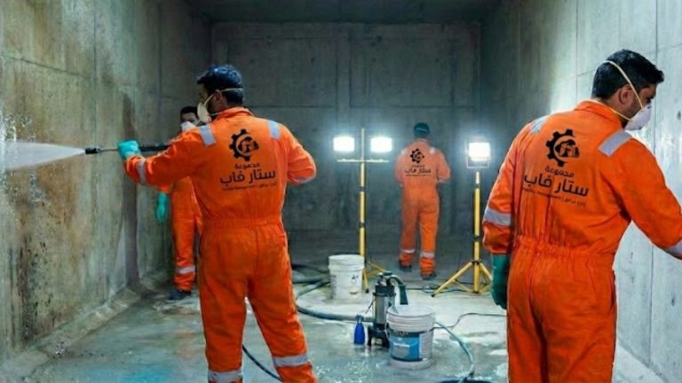 ثلاثة عمال من شركة ستار فاب يرتدون بدلات واقية برتقالية وكمامات، داخل خزان مياه خرساني كبير. أحدهم يستخدم مضخة مياه عالية الضغط لغسيل الجدران، والآخرون يجهزون المعدات تحت إضاءة كشافات قوية.